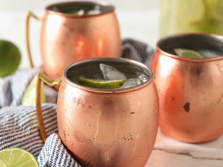 Three copper mugs sitting on a blue and white striped towel hold Moscow mules garnished with lime wedges on marble with a pitcher behind them
