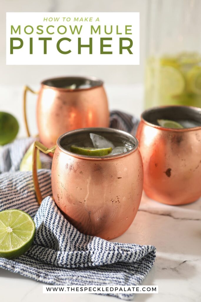 Three copper mugs sitting on a blue and white striped towel hold Moscow mules garnished with lime wedges on marble with a pitcher behind them with the text 'how to make a moscow mule pitcher'