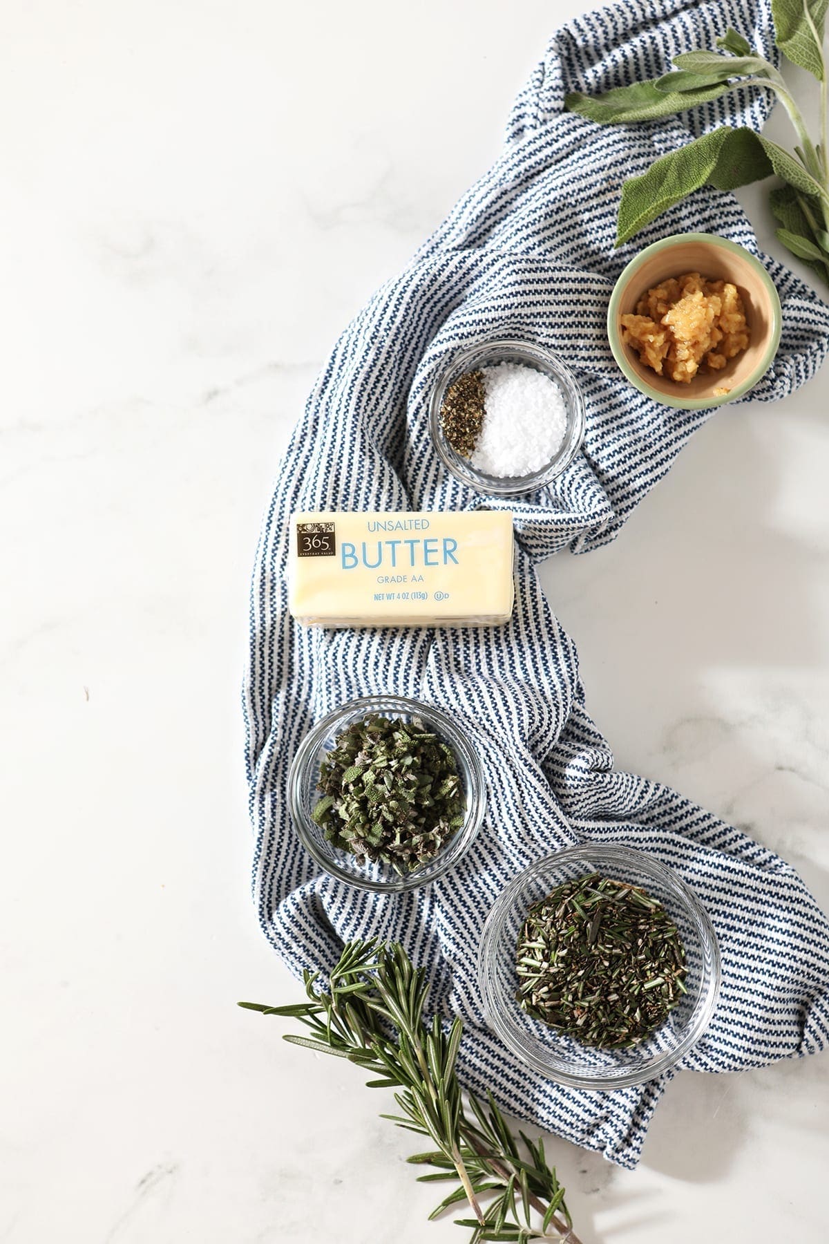 Butter, herbs, salt and pepper and garlic in separate bowls on a blue striped towel