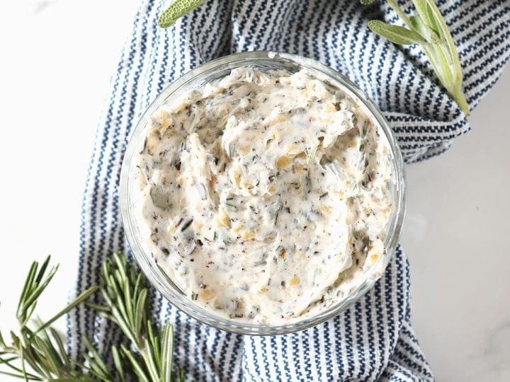 Blended herb butter in a glass bowl on a blue striped towel surrounded by fresh sage and rosemary