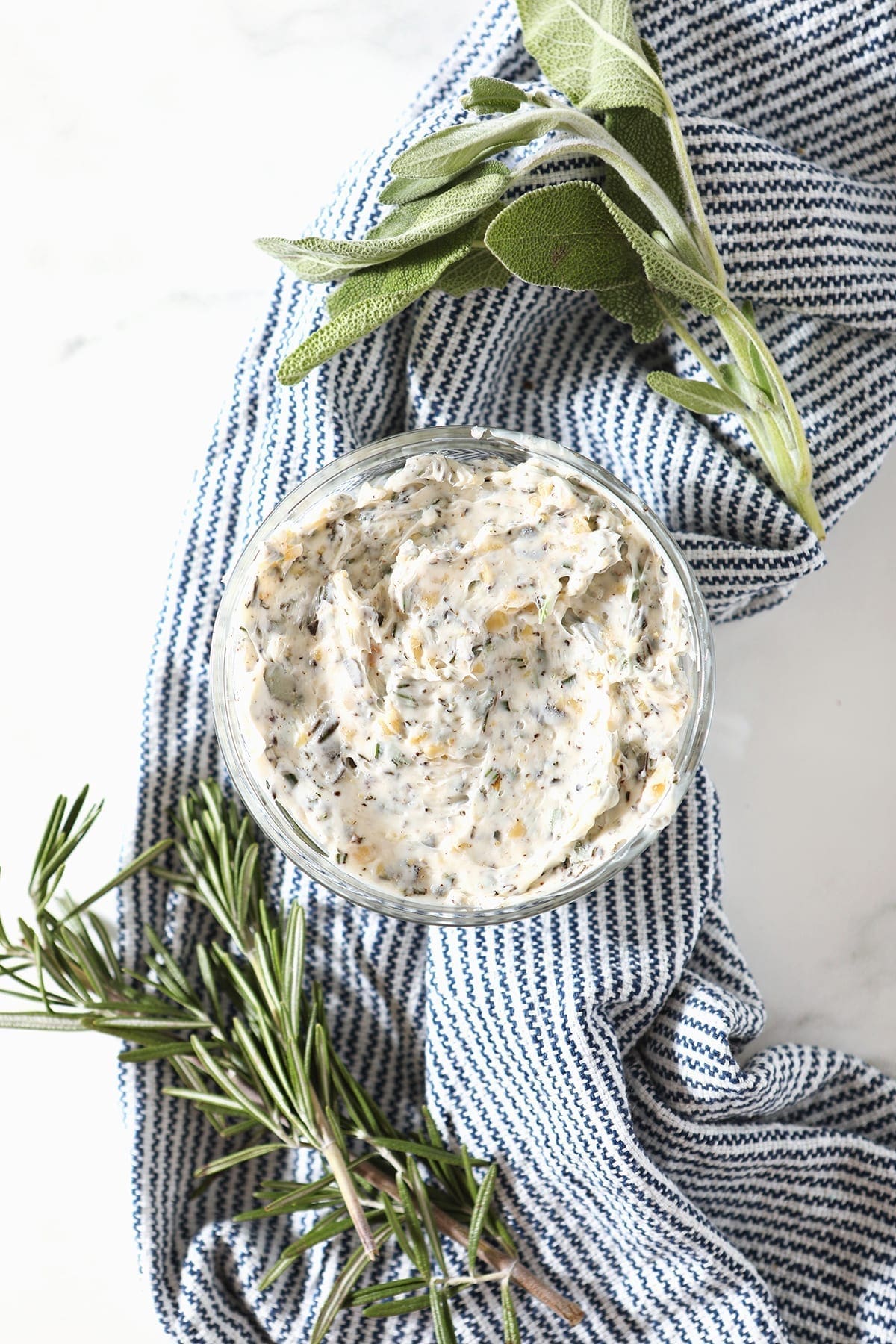 Blended herb butter in a glass bowl on a blue striped towel surrounded by fresh sage and rosemary