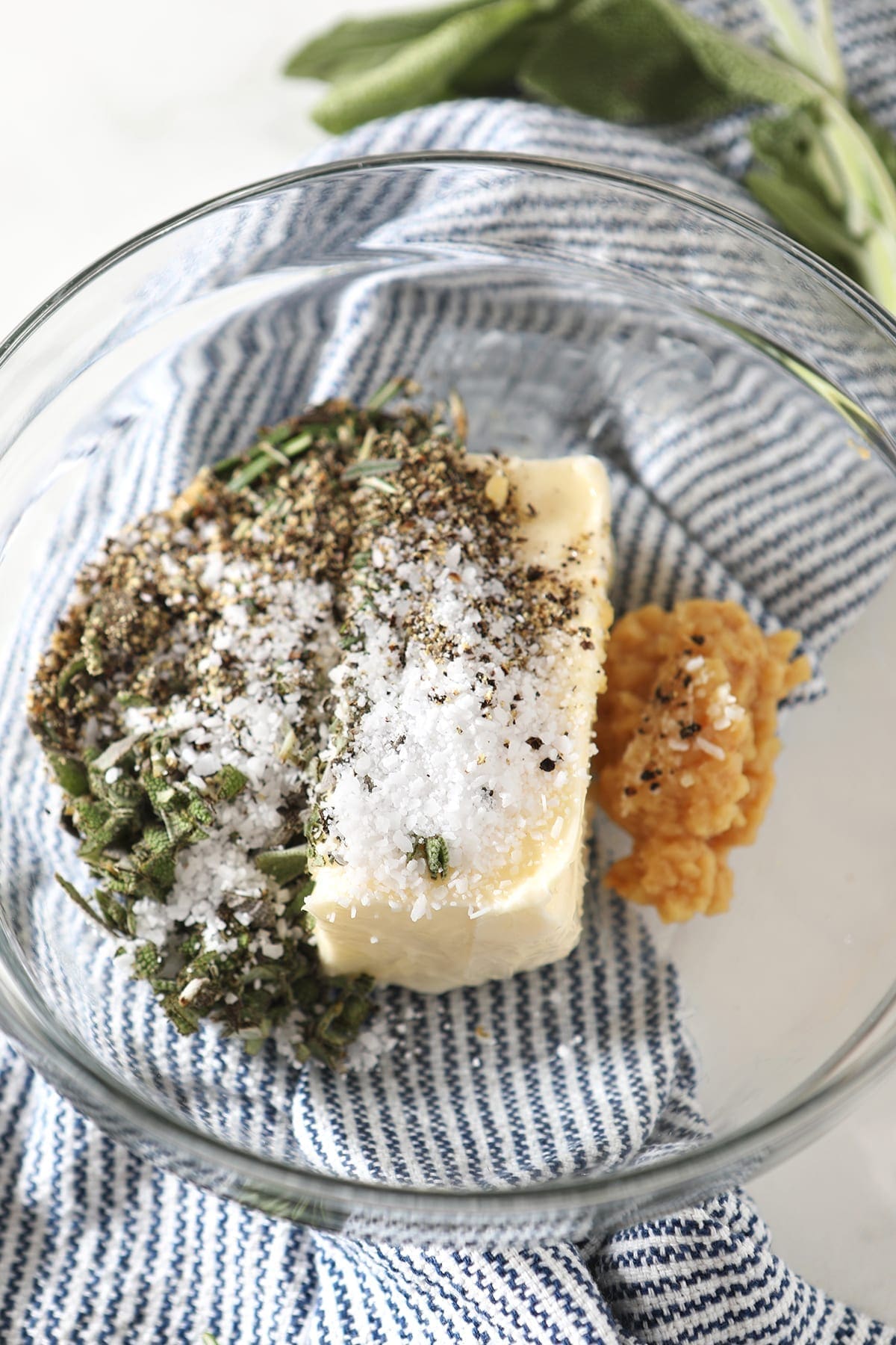 Herb butter ingredients in a glass bowl sitting on a blue striped towel before blending