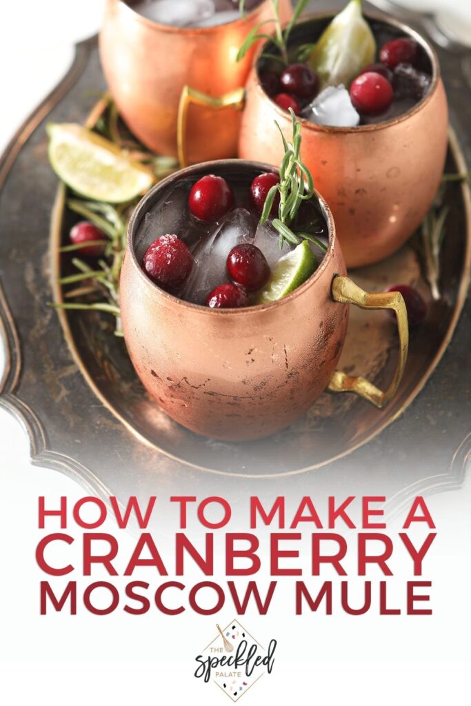 Three Yule Mules, garnished with fresh cranberries, lime wedges and rosemary sprigs, sit on a silver platter with the text 'how to make a cranberry moscow mule'