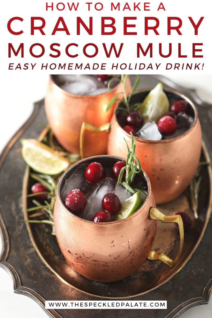 Three Yule Mules, garnished with fresh cranberries, lime wedges and rosemary sprigs, sit on a silver platter with the text 'how to make a cranberry moscow mule. easy homemade holiday drink!'