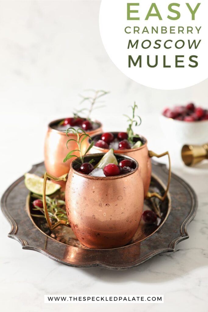 Three Yule Mules, garnished with fresh cranberries, lime wedges and rosemary sprigs, sit on a silver platter with the text 'easy cranberry moscow mules'