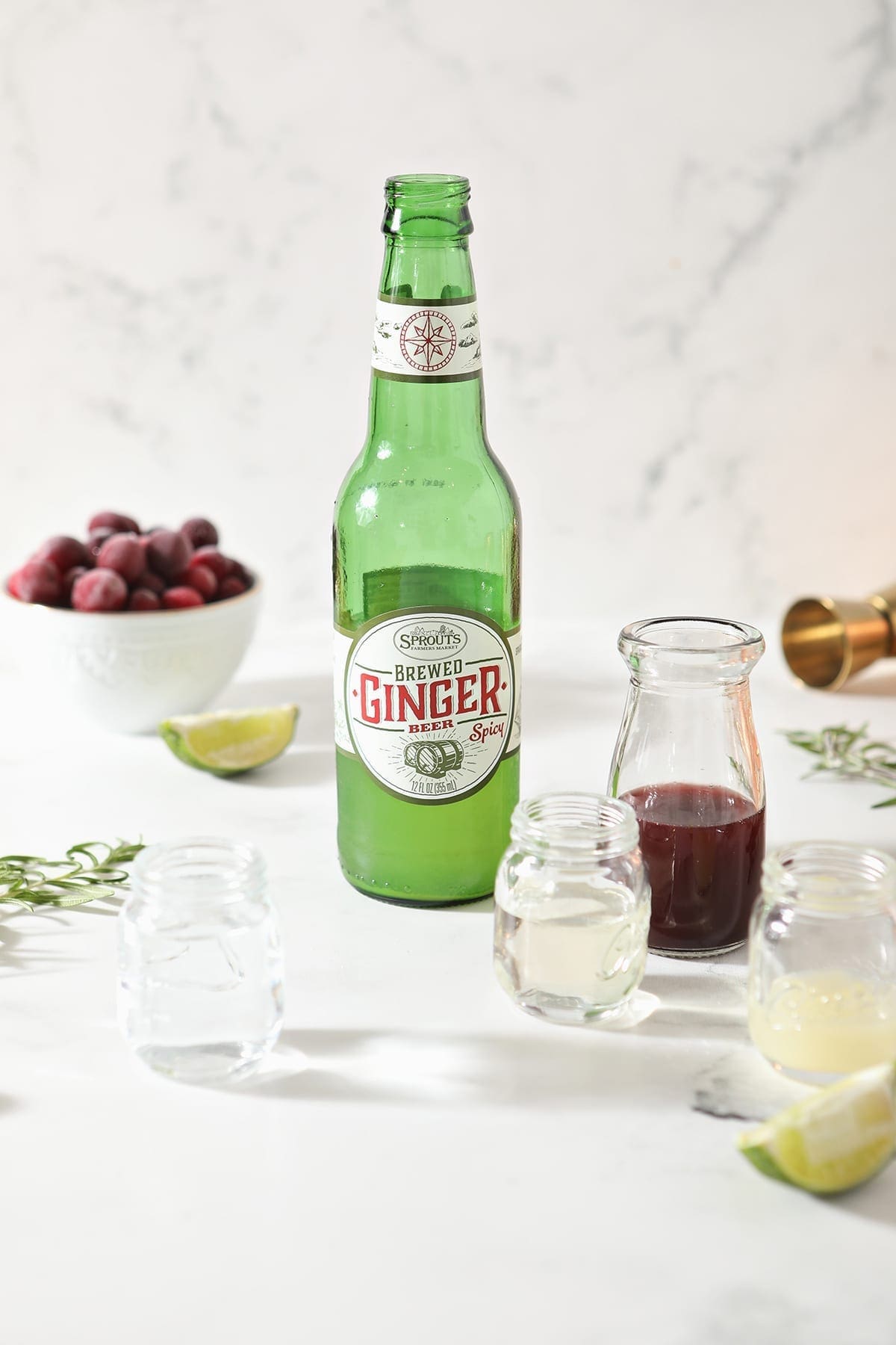 Ginger beer in its bottle sits next to shot glasses holding lime juice, vodka, simple syrup and lime juice on marble