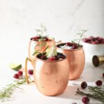 Three Cranberry Moscow Mules in copper cups sit on marble next to sprigs of rosemary and fresh cranberries