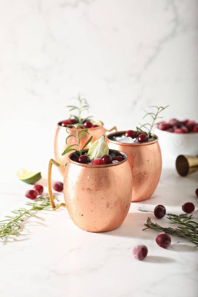 Three Cranberry Moscow Mules in copper cups sit on marble next to sprigs of rosemary and fresh cranberries