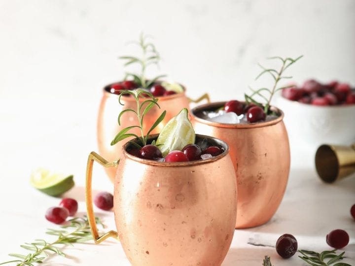 Three Cranberry Moscow Mules in copper cups sit on marble next to sprigs of rosemary and fresh cranberries