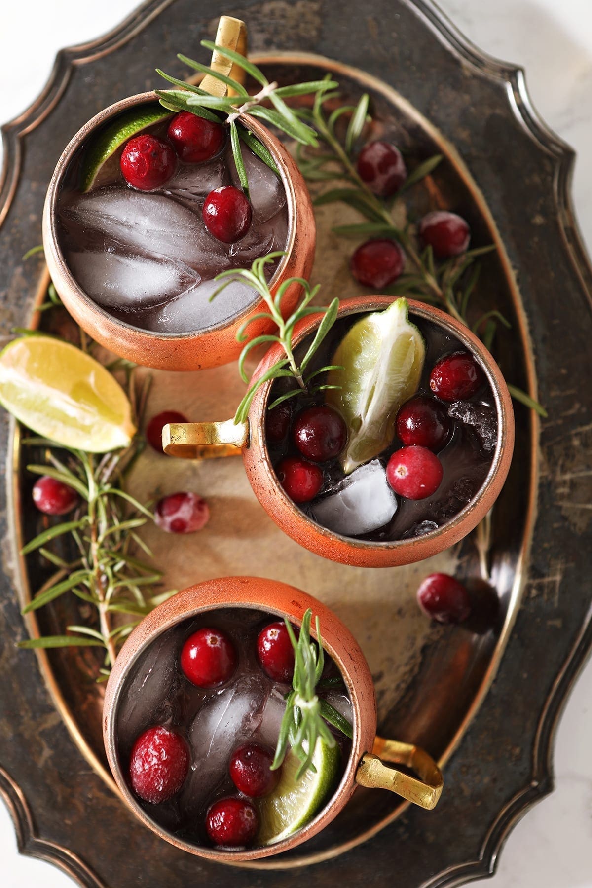 Three Yule Mules, garnished with fresh cranberries, lime wedges and rosemary sprigs, sit on a silver platter