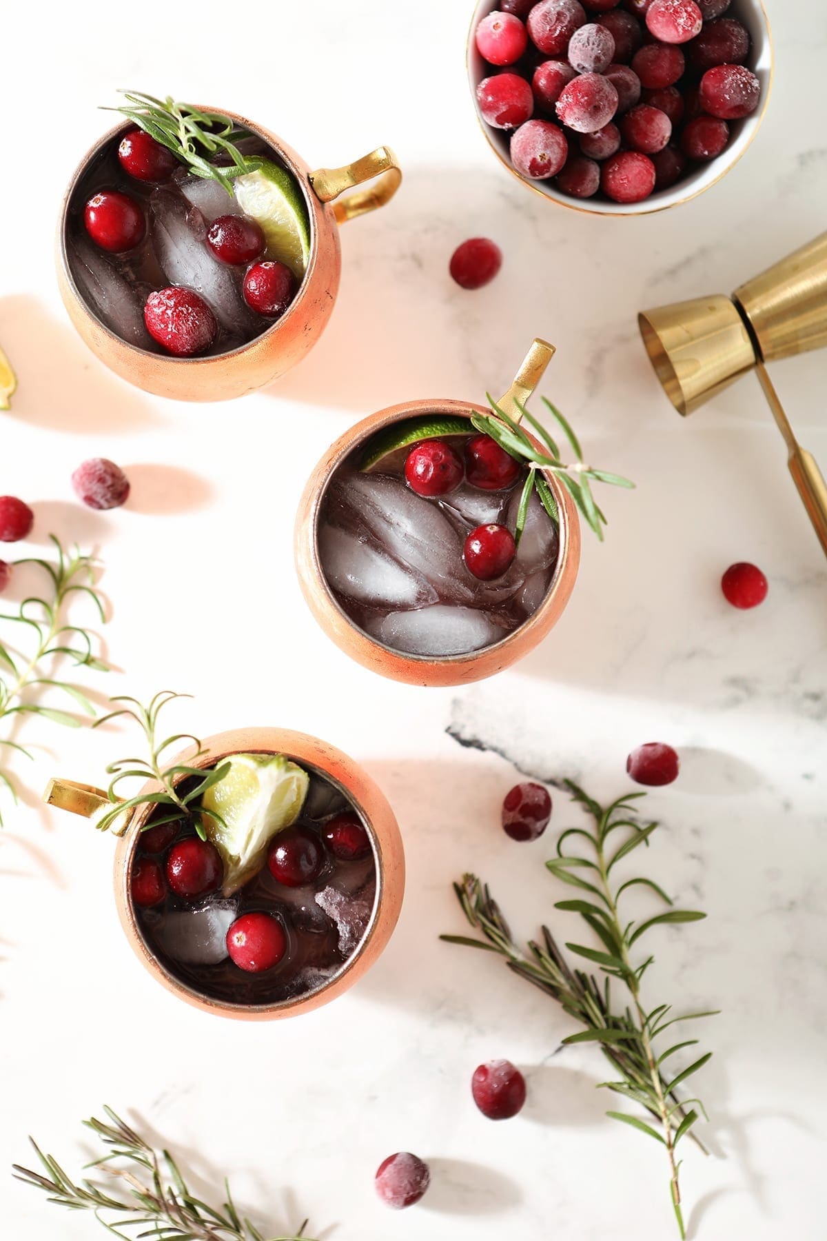 Three Cranberry Mules sit on a marble surface next to a bowl of cranberries, a gold jigger, rosemary sprigs and more cranberries