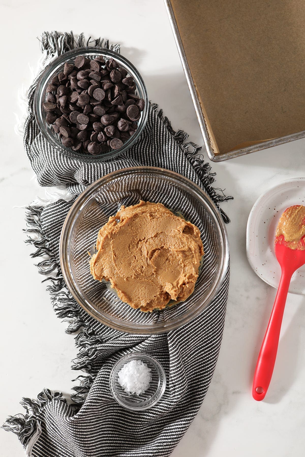 A bowl of peanut butter with a bowl of chocolate chips and flaky sea salt on top of a gray and white striped towel