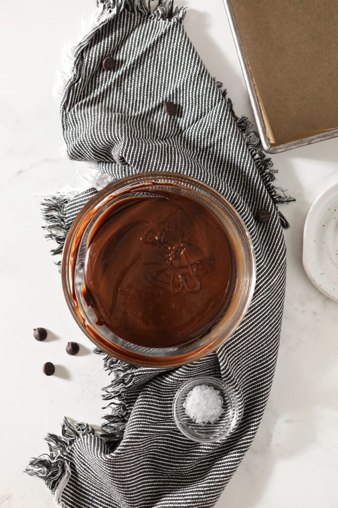 The melted peanut butter and chocolate mixture in a bowl on top of a gray and white towel next to a bowl of flaky sea salt and a baking dish