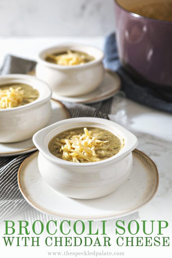 White bowls on pottery plates hold broccoli soup, garnished with extra cheese, on top of gray striped towels and next to a purple Dutch oven with the text 'broccoli soup with cheddar cheese'