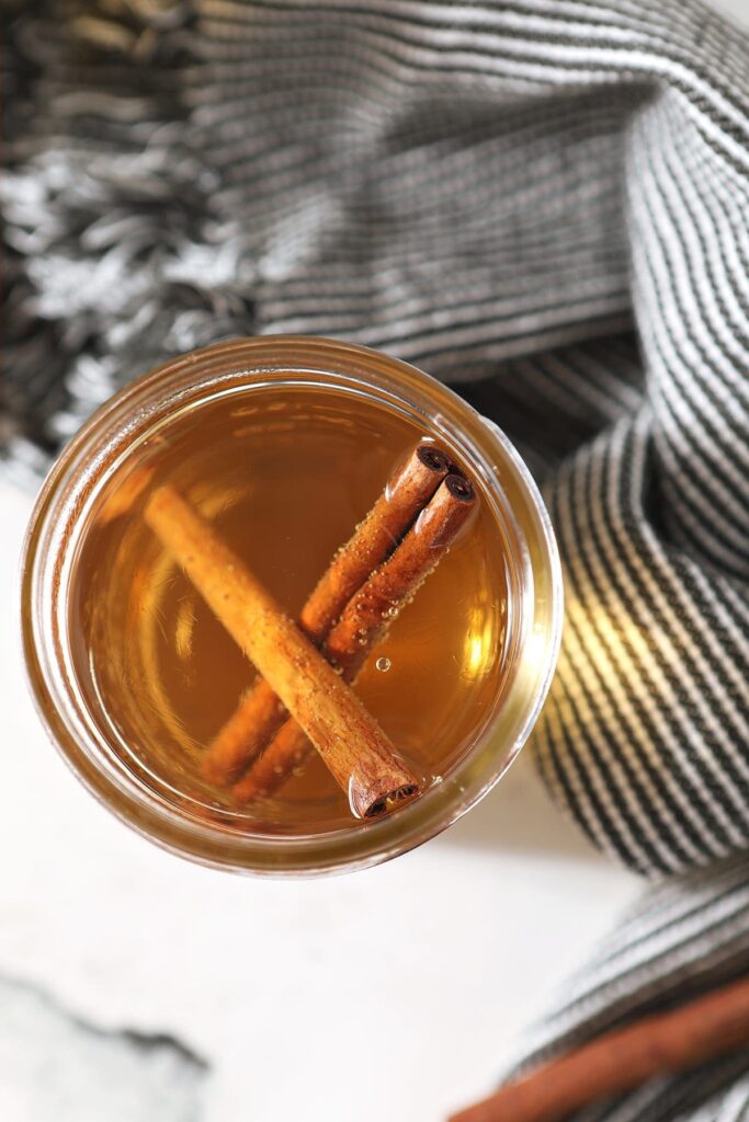 A jar of cinnamon simple syrup sits next to a gray and white striped towel with cinnamon sticks inside it