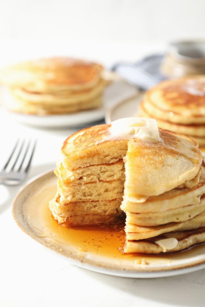 A stack of old fashioned homemade pancakes on a white plate with syrup and butter with a wedge cut out of it