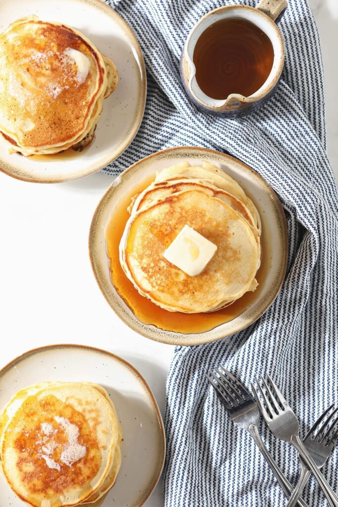 Three plates holding stacks of pancakes topped with butter sit on a blue striped towel next to forks and a container of syrup