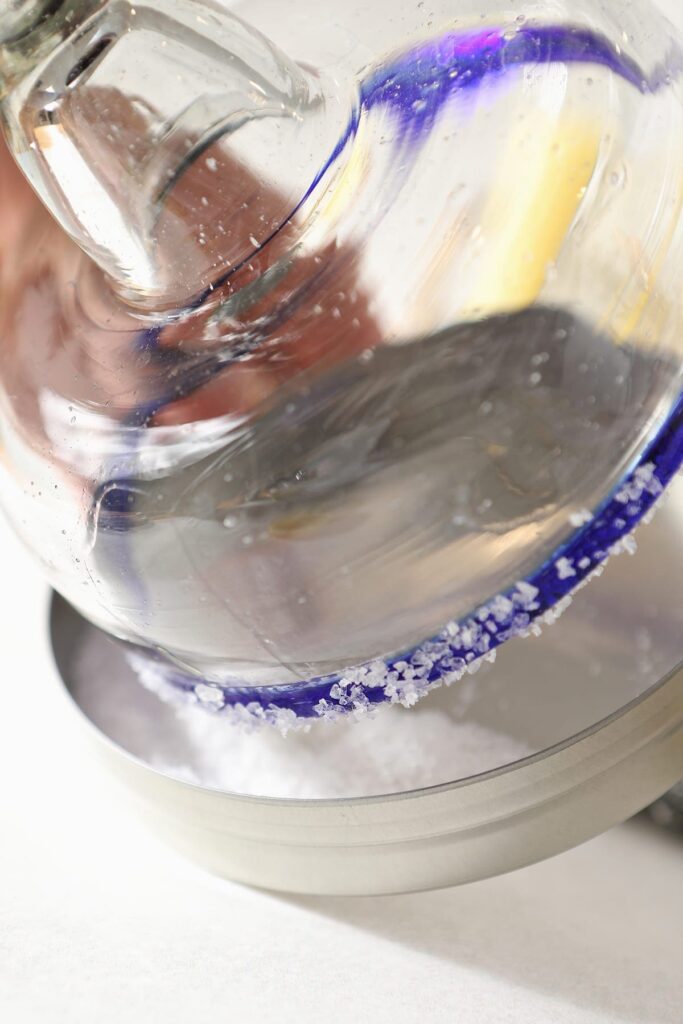 A blue-rimmed margarita glass is dipped into a metal container holding salt