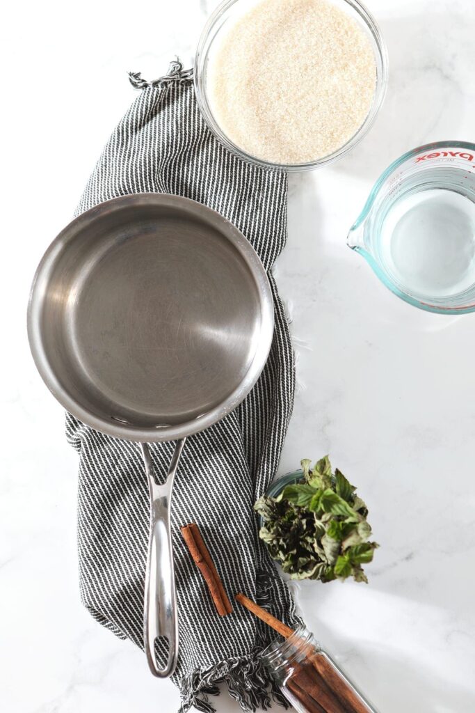 A saucepan sits on a striped towel next to cinnamon sticks, a bunch of mint, sugar and water