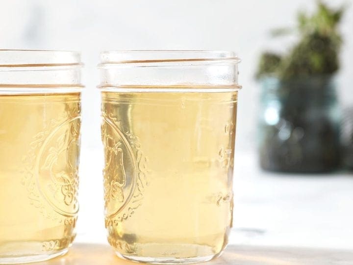 Two jars holding simple syrup sit on a marble surface in front of a jar holding fresh mint leaves