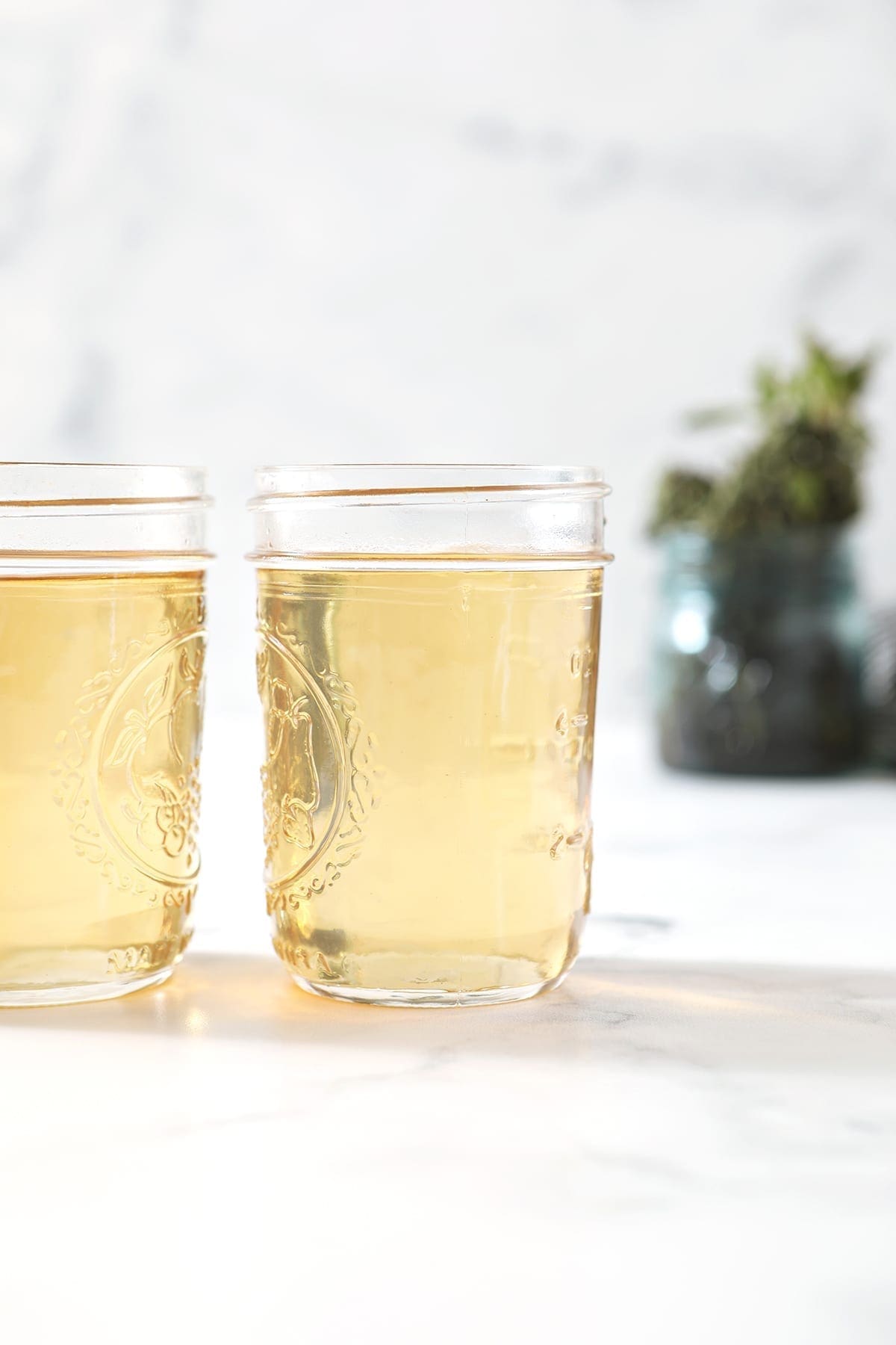 Two jars holding simple syrup sit on a marble surface in front of a jar holding fresh mint leaves