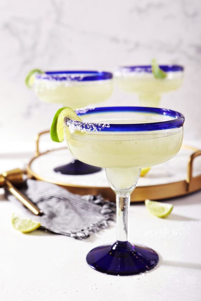 A salt-rimmed glass holding a classic margarita recipe sits on marble, in front of a marble tray holding two more