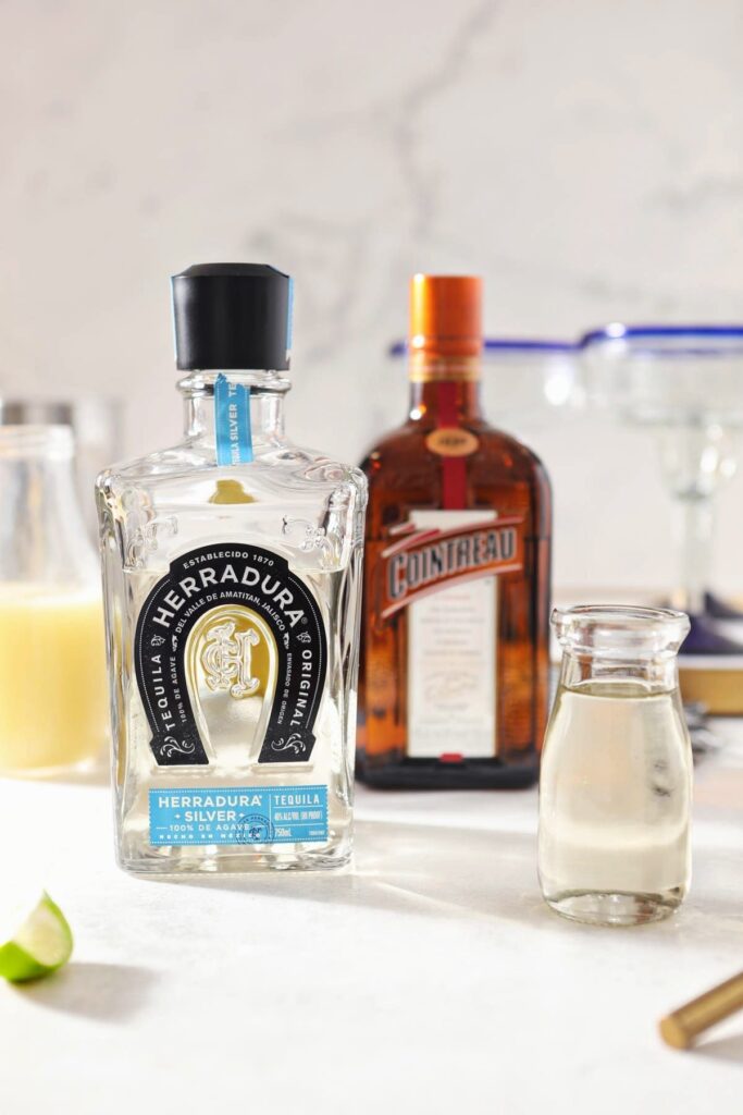 A bottle of tequila sits next to lime juice, simple syrup and a bottle of Countreau on a marble counter