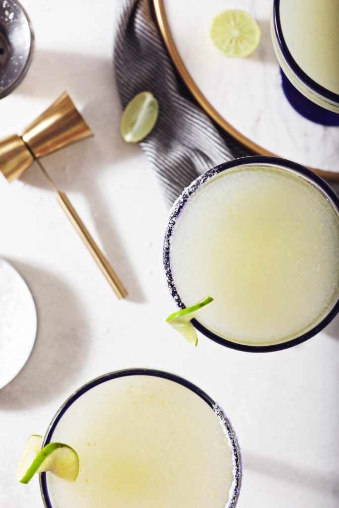 Three Cointreau margaritas sit on a marble counter next to a jigger, lime wedges and a container of salt