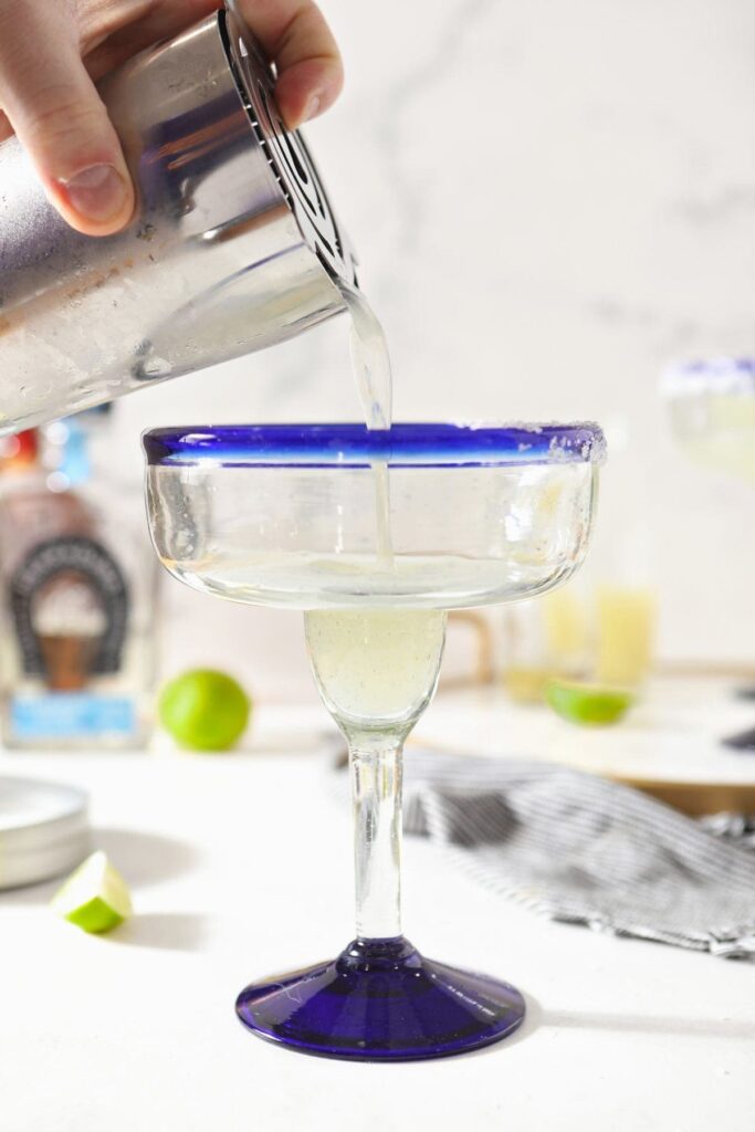 A person pours a drink into a salt-rimmed blue margarita glass