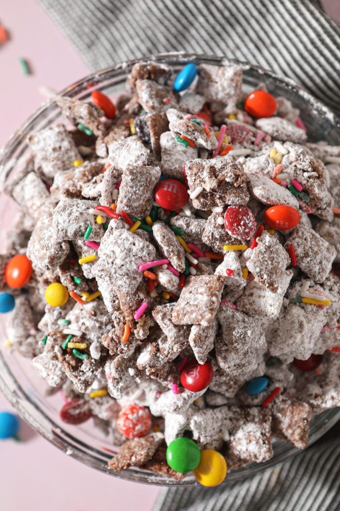 Close up of a bowl with rainbow sprinkles and chocolate-covered candies in Chex Mix Puppy Chow