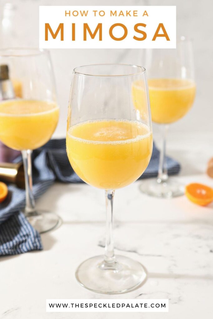 A prosecco mimosa in a wine glass sits in front of two more on a marble countertop