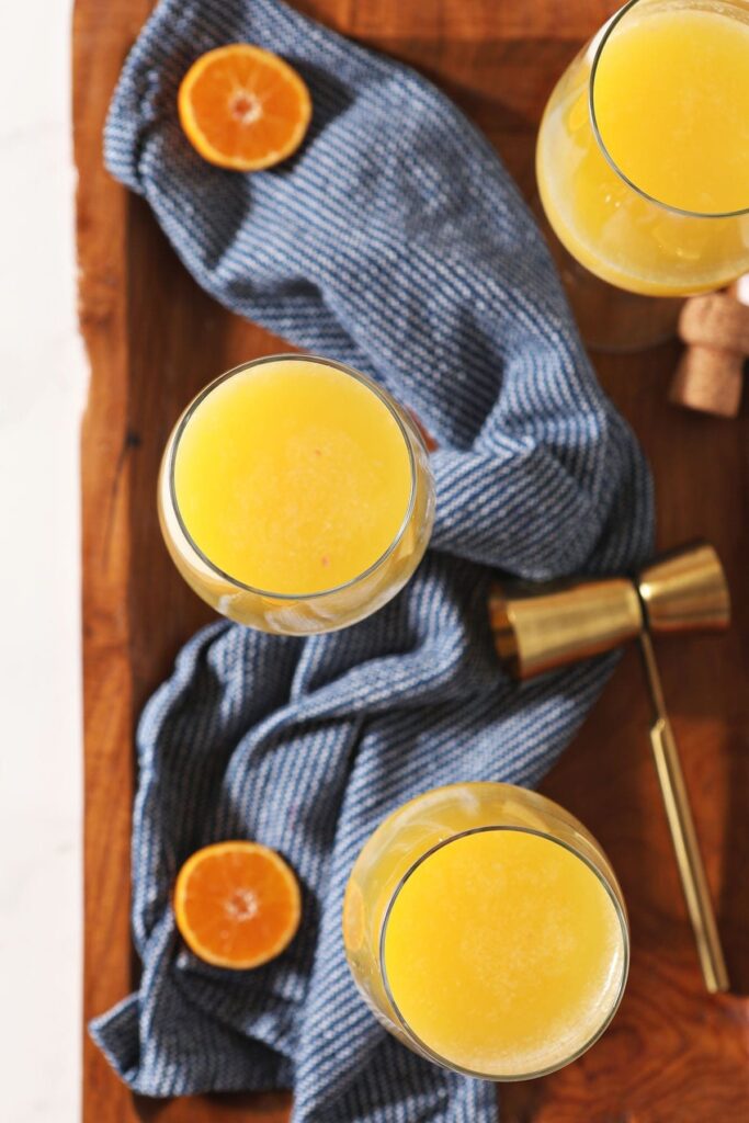 Three wine glasses holding orange juice and champagne sit on a wooden tray