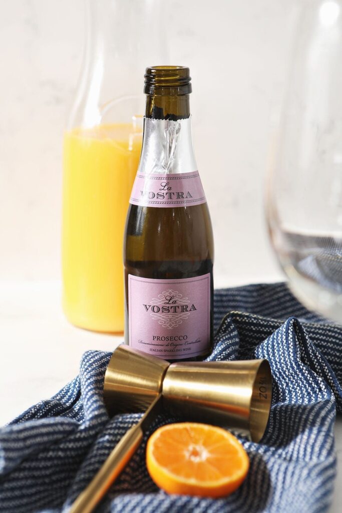 A small bottle of prosecco with a jigger, a bottle of orange juice and an orange round on a dark blue linen