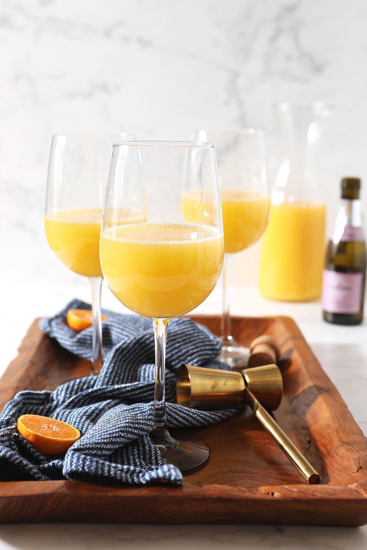 A small bottle of prosecco with a jigger, a bottle of orange juice and an orange round on a dark blue linen