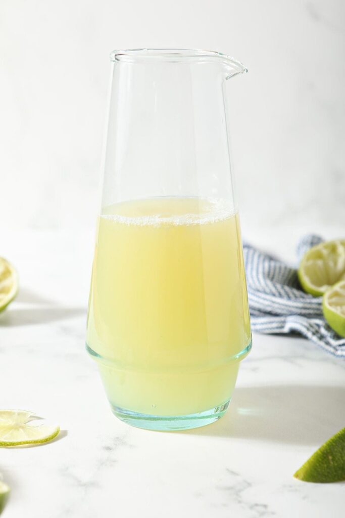 A pitcher of margarita mix sits in front of a blue and white striped towel surrounded by limes