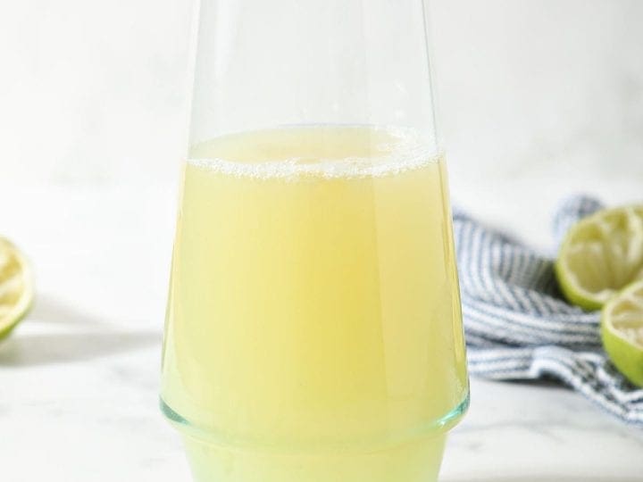 A pitcher of margarita mix sits in front of a blue and white striped towel surrounded by limes