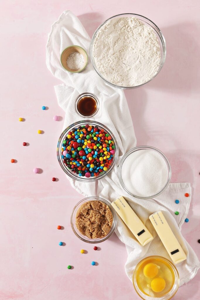 Ingredients to make M&M sugar cookies in bowls sit on a white towel on top of a pink countertop