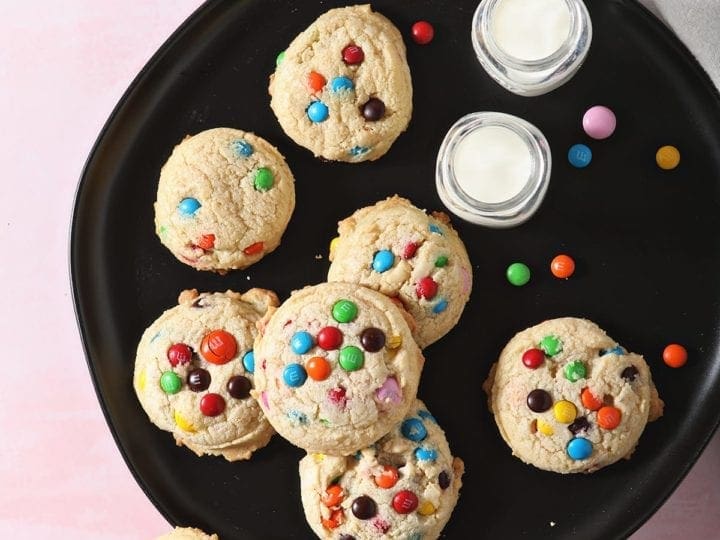 M&M Cookies sit on a black plate next to glasses of milk on a pink countertop