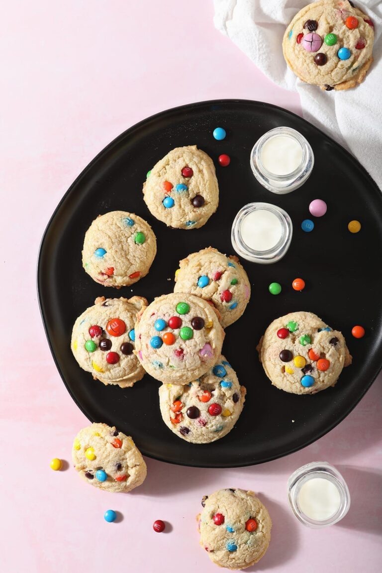 M&M Cookies sit on a black plate next to glasses of milk on a pink countertop