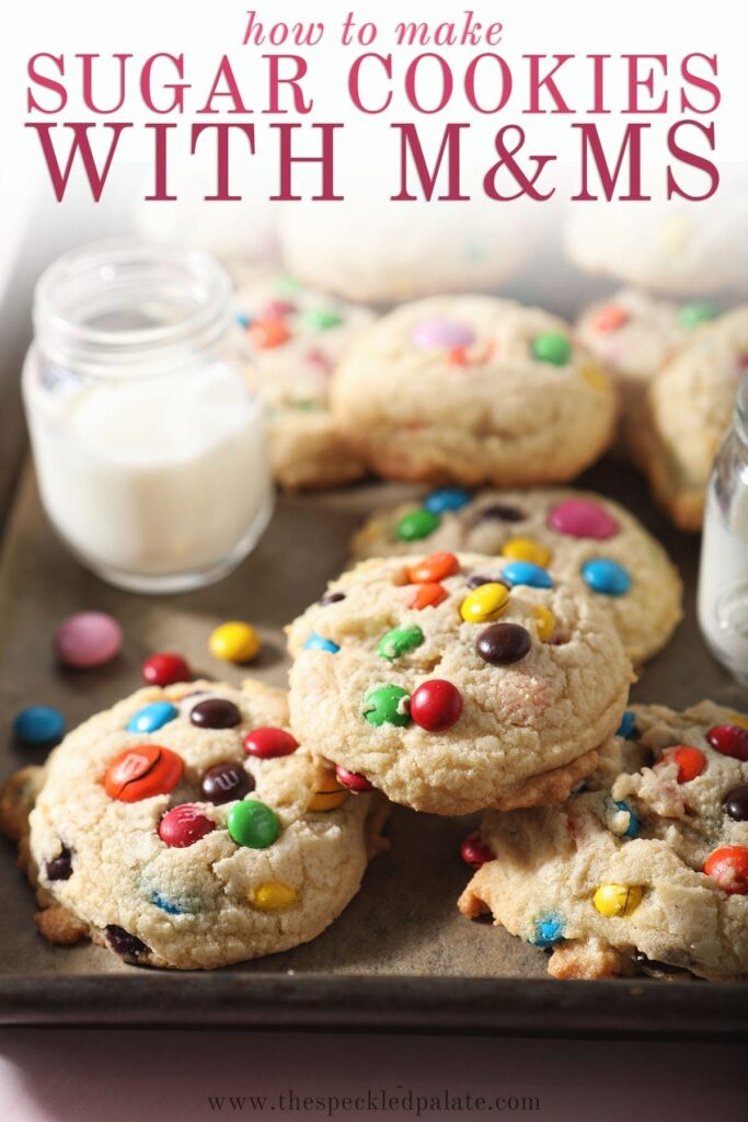 A baking sheet holds several M&M cookies next to a glass of milk with the text 'how to make sugar cookies with m&ms'