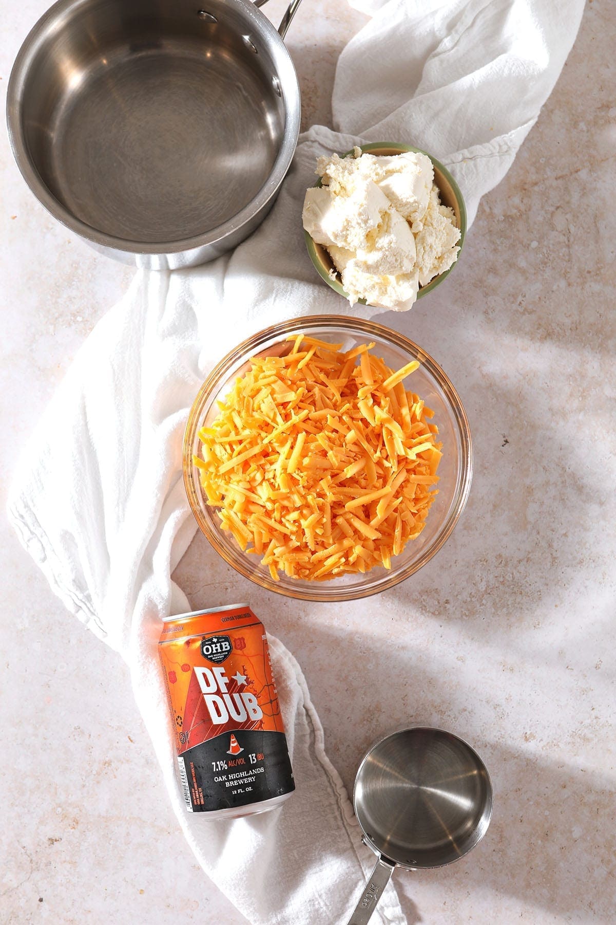 Ingredients for beer cheese dip sit on a countertop next to a measuring cup and a small saucepan