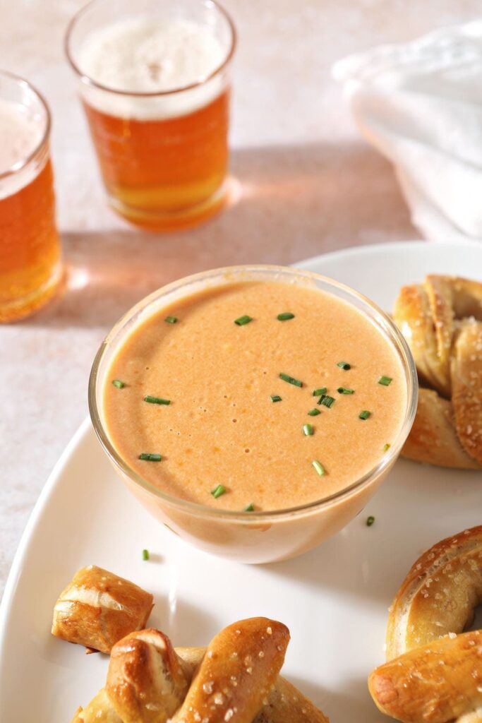 A bowl holding Pretzel Cheese Dip sits on a white tray next to pretzels and near two cups of beer