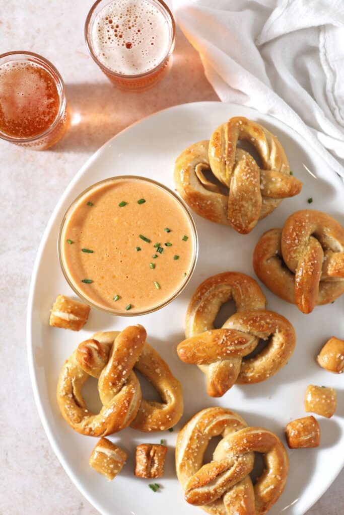 Pretzel Cheese Dip in a bowl sits on a white plate with pretzels and pretzel bites next to beer glasses