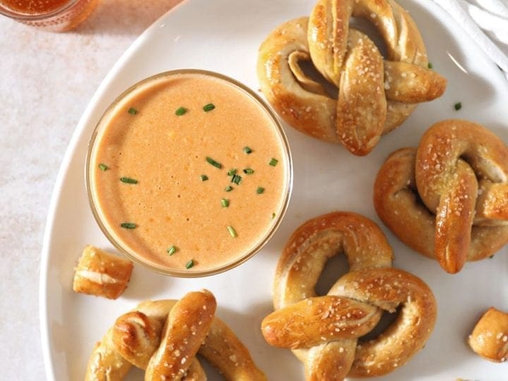 Pretzel Cheese Dip in a bowl sits on a white plate with pretzels and pretzel bites next to beer glasses