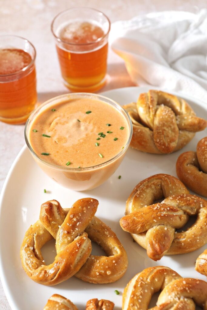 Homemade soft pretzels sit on a white tray next to a bowl of cheese sauce for pretzels and beer