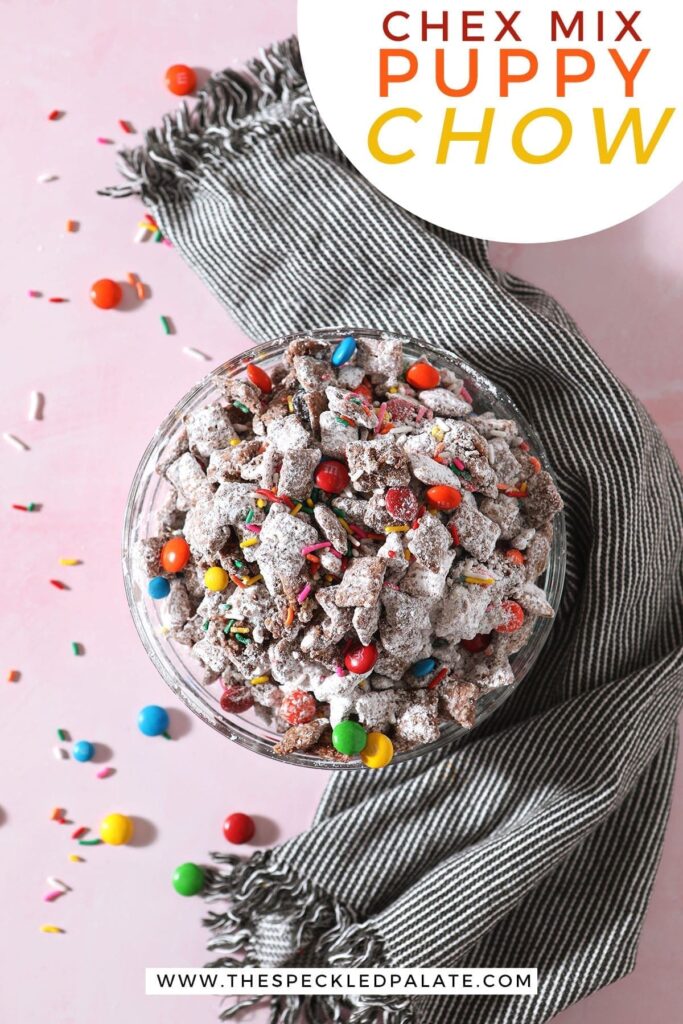 A bowl of rainbow puppy chow sits on a pink countertop next to a gray striped towel with the text 'chex mix puppy chow'