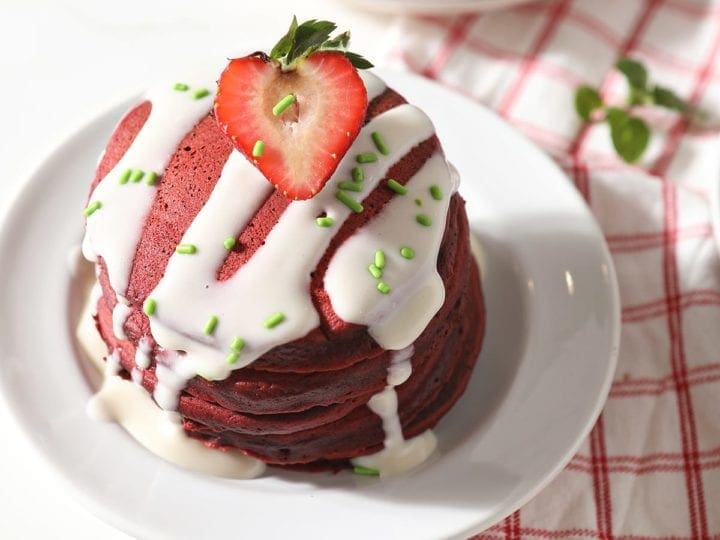 A stack of red velvet pancakes is garnished with a cream cheese glaze, green sprinkles and a halved strawberry on a white plate