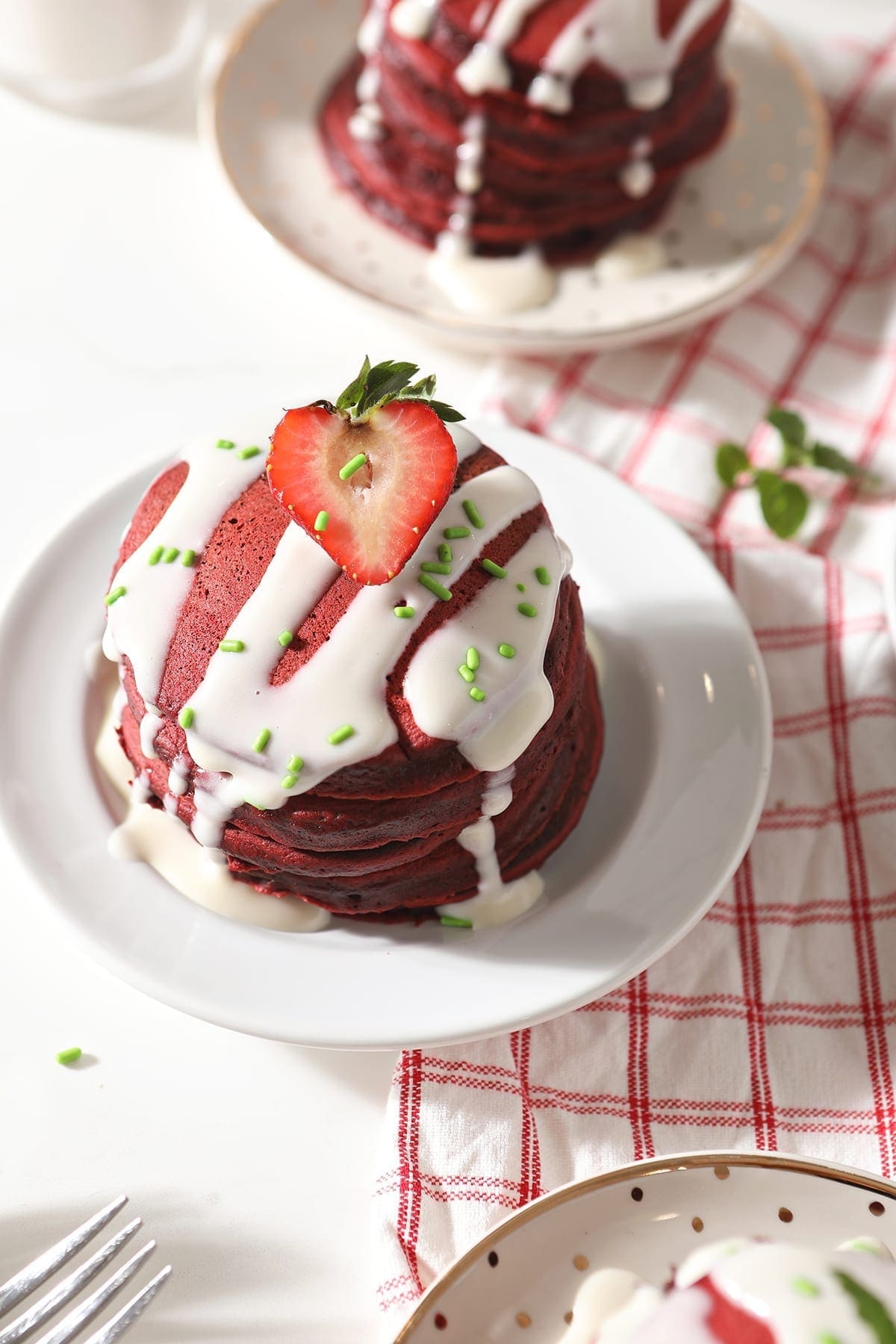 A stack of red velvet pancakes is garnished with a cream cheese glaze, green sprinkles and a halved strawberry on a white plate