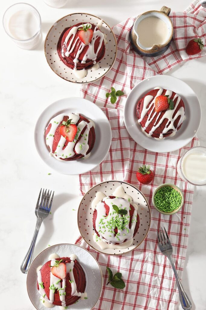 Five plates holding garnished red pancakes sit on a red and white towel on a marble countertop