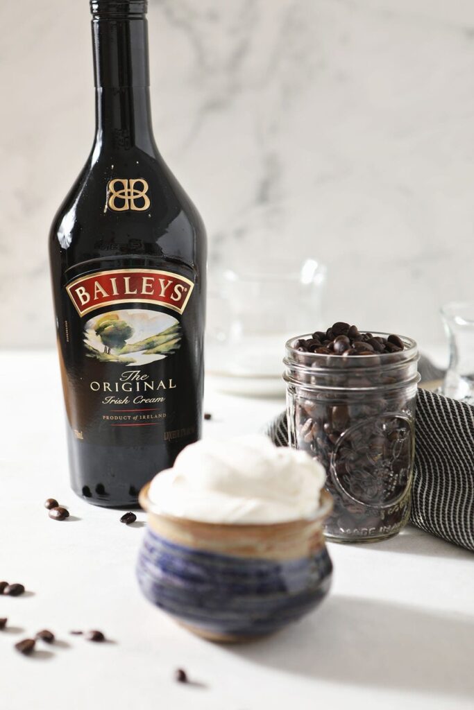 A bottle of Irish Cream, a jar of coffee beans and a bowl of whipped cream sit together on a marble countertop